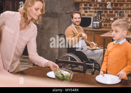 Mère avec fils table de service pour le dîner tandis que le père à couper des légumes en fauteuil roulant à la maison Banque D'Images