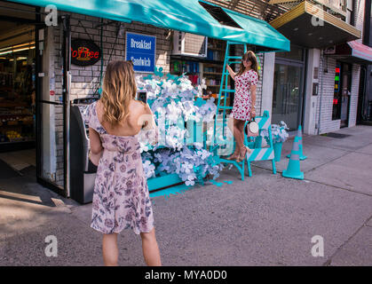 Young woman photographing son ami debout sur l'échelle, à l'extérieur de l'atelier, les deux femmes portant des robes à motifs floraux, New York City, USA Banque D'Images