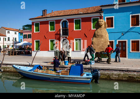 Les femmes achètent des fruits et légumes frais d'un magasin mobile comme un groupe d'hommes poussent un carnaval flotter le long de la rue, l'île de Burano, Venise, Italie Banque D'Images