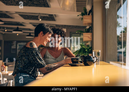 Female friends sitting at coffee shop et souriant. Young woman using mobile phone alors qu'il était assis avec son amie au coffee shop. Banque D'Images