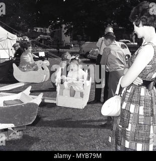 1963, historiques, de la mère à l'extérieur sur l'herbe par sa petite fille qui est sur un terrain de merry-go-round ride à une fête de charité, England, UK. Banque D'Images