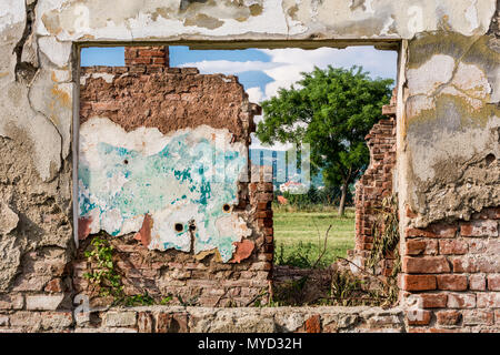 Mur endommagé et le cadre de la fenêtre avec vue sur les ruines de prix et terrain vert d'herbe, de l'arbre et le bleu ciel nuageux. Mur de brique rouge avec des fissures Banque D'Images