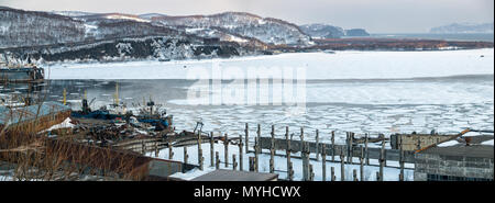 Vue panoramique de l'ancien chantier naval, coques de production, de vieux bateaux, navires, baie couverte de glace, collines couvertes de neige, des rochers dans la distance en avril après Banque D'Images