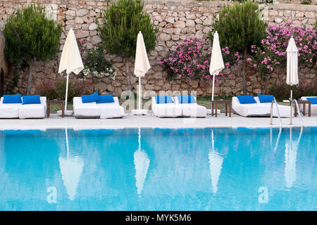 Une piscine vide avec chaises longues blanches, coussins bleus et parasols, reflétant l'eau calme, Banque D'Images