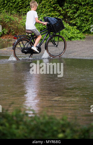 Young boy riding a bike à travers l'eau d'inondation dans une rue après la pluie torrentielle Banque D'Images