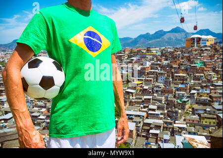 Joueur de football au Brésil permanent flag t-shirt holding soccer ball en face de favela arrière-plan des bidonvilles de Rio de Janeiro. Banque D'Images