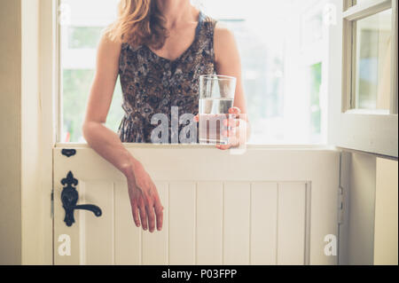 Une jeune femme se tient debout par une porte de l'écurie avec un verre d'eau dans sa main Banque D'Images