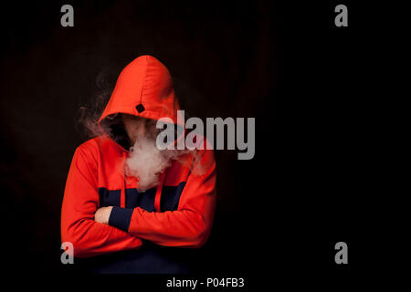 Un jeune homme dans un capot orange debout dans une posture fermée avec tête abaissée fume une vape et exhale une fumée blanche sur un fond noir isolé Banque D'Images