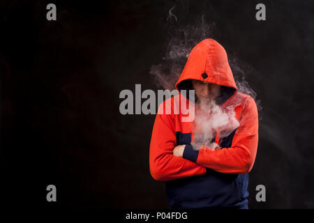 Un jeune homme dans un capot orange debout dans une posture fermée avec tête abaissée fume une vape et exhale une fumée blanche sur un fond noir isolé Banque D'Images