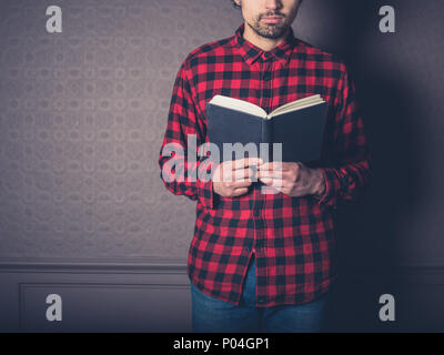 Un jeune homme portant une chemise de flanelle rouge est en train de lire un livre dans une salle de séjour de luxe Banque D'Images