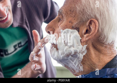 Un homme âgé se raser la barbe par jeune homme du métier à la maison. L'homme l'application de la mousse à l'ancien visage de l'homme. Banque D'Images