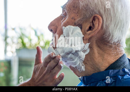 Un homme âgé se raser la barbe par jeune homme du métier à la maison. L'homme l'application de la mousse à l'ancien visage de l'homme. Banque D'Images