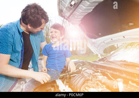 Portrait d'un garçon de 7 ans assis sur le capot de la voiture ouvert cassé tandis que son père la réparation à l'extérieur du moteur Banque D'Images