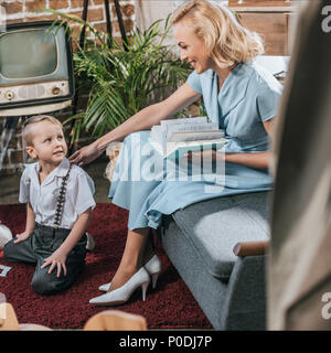 Happy blonde woman reading book et à la recherche au petit fils de jouer sur un tapis à la maison familiale de style années 1950, Banque D'Images