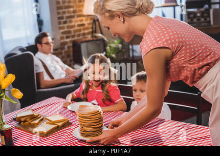 Enfants heureux à la recherche à smiling mother putting crêpes sur la table, de style rétro Banque D'Images