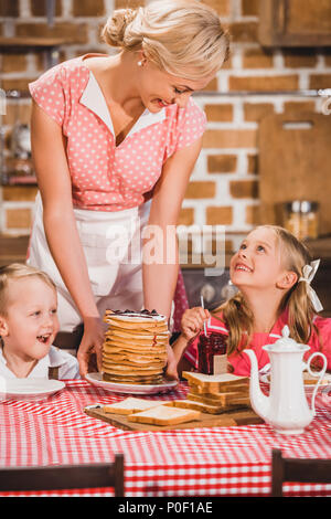 Happy mother putting crêpes sur la table et regardant cute smiling kids le petit-déjeuner, 50s style famille Banque D'Images