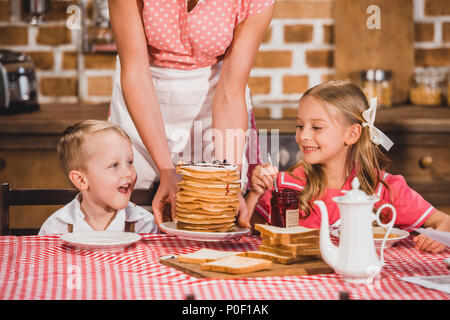 Cropped shot of mother putting de crêpes sur la table et regardant cute smiling kids le petit-déjeuner, 50s style famille Banque D'Images