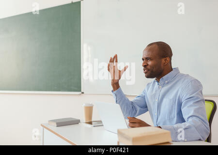 Frustrés african american teacher gesticulant et sitting at desk with laptop Banque D'Images