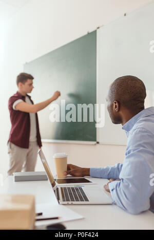 High school student écrit au tableau et african american teacher sitting at desk with laptop and Coffee cup Banque D'Images