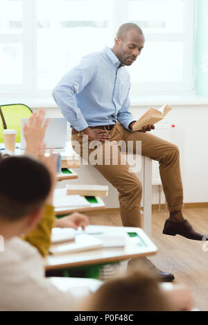 African American teacher sitting on table et livre de lecture aux enfants des écoles avec les bras en classe Banque D'Images