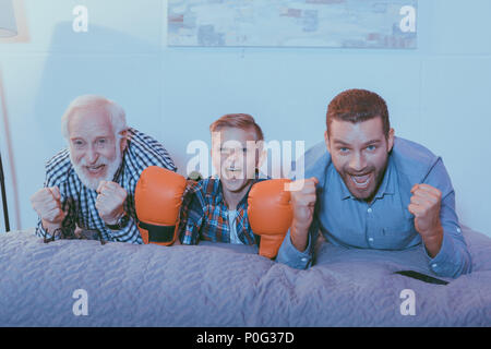 Little Boy wearing boxing gloves, son père et son grand-père étendu sur le lit, regardant un match de boxe et acclamer Banque D'Images