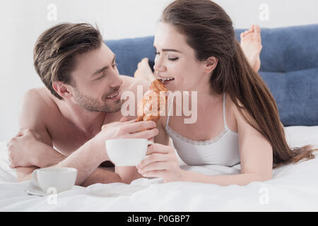 Beau jeune homme se nourrir sa petite amie au lit avec croissant et café Banque D'Images