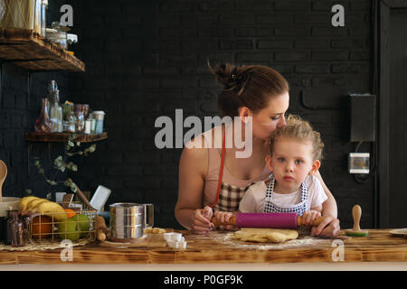 Jeune mère et fille préparer des cookies dans la cuisine. Ils sont dans des tabliers. Petite fille roule la pâte avec un rouleau à pâtisserie. Bisous femme enfant, encourageant les di Banque D'Images