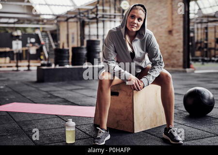 Souriante jeune femme blonde en vêtements d'exercice assis seul sur une case dans un gymnase d'entraînement de penser à elle Banque D'Images