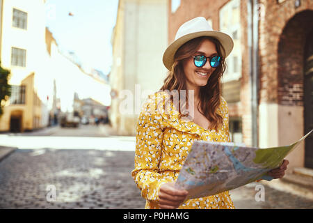 Souriante jeune femme brune portant un chapeau et des lunettes à explorer la ville avec une carte Banque D'Images