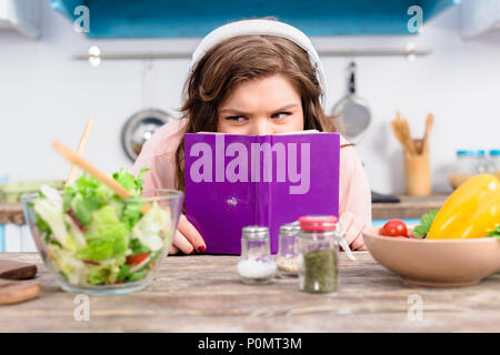 Vue de l'excès de femme masquée dans les écouteurs se cacher derrière livre à table avec une salade fraîche dans la cuisine à la maison Banque D'Images