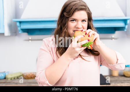 Portrait de jeune femme en surpoids manger burger dans les mains dans la cuisine à la maison Banque D'Images