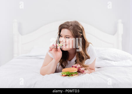 Femme en surpoids manger pyjama burger on bed at home Banque D'Images