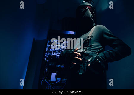 Low angle view of hacker dans les vêtements noirs et lunettes avec différents câbles sur l'arrière-plan Banque D'Images