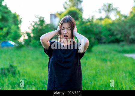 Jeune fille brune en dehors du parc. Mains tenant les oreilles. Il a crié très fort. Son fort solide. Il est impossible d'écouter le bruit. Ses yeux pressés émotionnellement ouvrir légèrement la bouche. Des vêtements décontractés. Banque D'Images