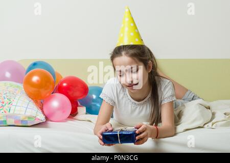 L'anniversaire d'une adolescente est de 10-11 ans. Une fille dans un chapeau de fête avec un cadeau se trouve sur le lit dans une chambre d'enfants, des ballons d'arrière-plan Banque D'Images