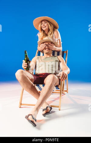 Smiling woman covering face of man holding beer en position assise sur une chaise sur fond bleu Banque D'Images