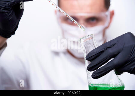 Une fiole avec un liquide vert et un tube à essai avec une goutte tombant dans les mains d'un scientifique de lunettes de protection, un masque et des gants noir isolé sur Banque D'Images