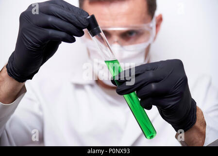 Mains d'un scientifique de gants noirs tenant un tube à essai avec un dangereux liquide chimique verte sur un fond blanc. Banque D'Images