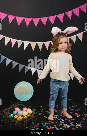 Beau petit enfant avec oreilles de lapin et oeufs de pâques en nid sur noir Banque D'Images