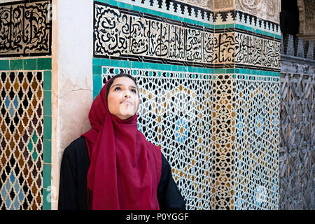 Femme musulmane dans des vêtements traditionnels avec hijab rouge et robe noire en face de arabesque traditionnel mur décoré avec du texte de Coran Banque D'Images