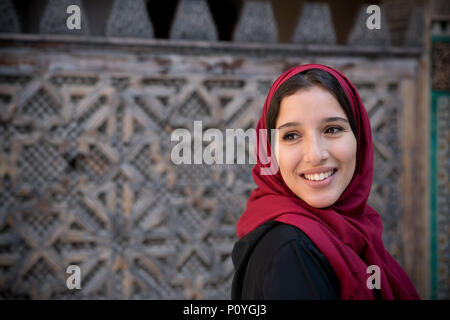 Portrait de femme musulmane smiling en costume traditionnel avec hijab rouge et robe noire en face de mur décoré arabesque traditionnels Banque D'Images