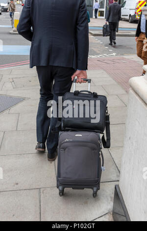 Un homme marchant le long d'une rue sur la chaussée le boulochage ou portant un wheelie cas et un porte-documents. homme portant des valises et des bagages sur pathway Banque D'Images