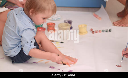 Cute little baby boy painting ses pieds sur un grand papier blanc vierge Banque D'Images