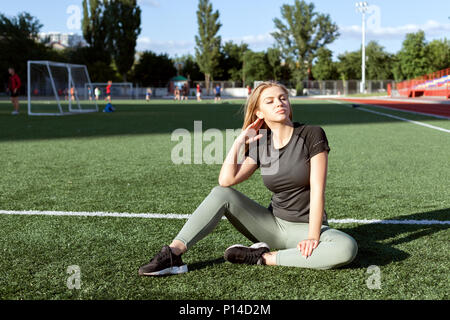 Jeune femme dans les vêtements de sport assis sur l'herbe au stade. Banque D'Images