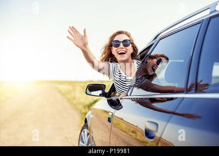 Jolie femme souriante en agitant sa main de la fenêtre de voiture un jour d'été. Banque D'Images