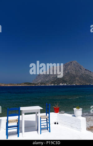 Table et chaises à l'île de Telendos dans la distance, Myrties, Kalymnos, îles du Dodécanèse, Grèce. Banque D'Images