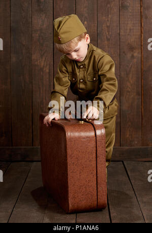 Les enfants garçon sont habillés en soldat en uniforme militaire rétro avec vieille valise, fond en bois foncé, style retro Banque D'Images