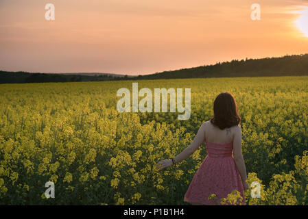 Jolie fille brune en robe rose, se promenant dans une culture de colza jaune pour le coucher du soleil la lumière, en Moravie du Sud, région de la République tchèque. Banque D'Images
