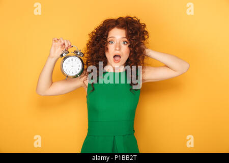 Portrait d'une femme rousse choqué dans dress holding réveil isolé sur fond jaune Banque D'Images