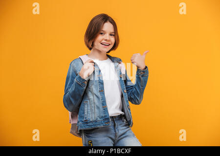 Portrait of a smiling little schoolgirl avec sac à dos à la caméra et au doigt pointant sur fond jaune Banque D'Images